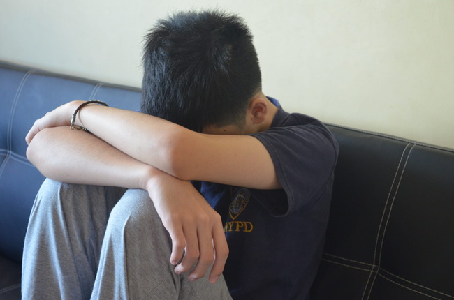 Teenage boy wearing grey sweatpants sitting with head on arms on black leather couch.